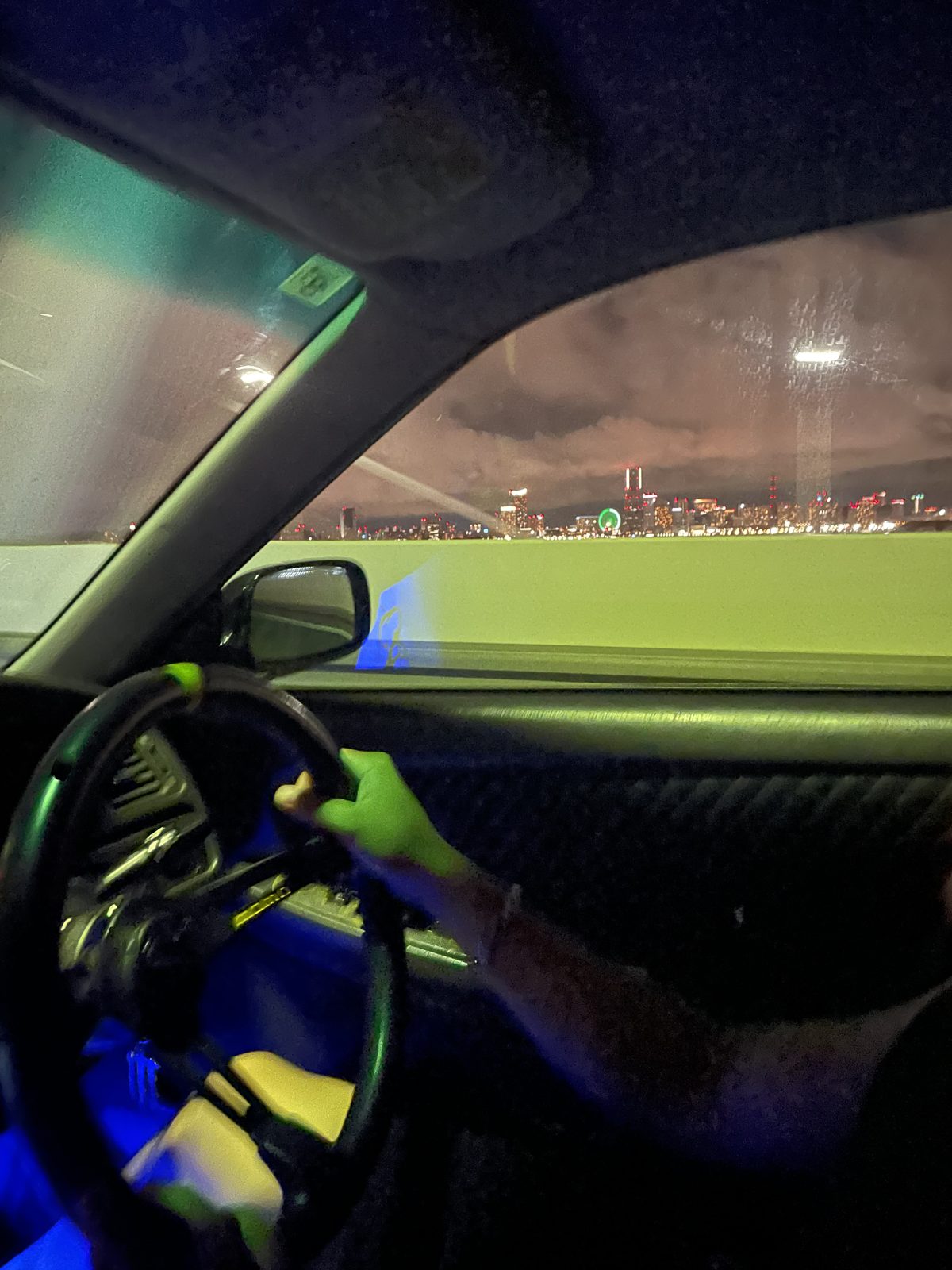 The Yokohama skyline at night seen through a car window from the expressway, with the driver's hand on the steering wheel